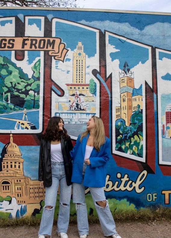 Two girls laughing in front of the Greeting from Austin Mural on South Congress