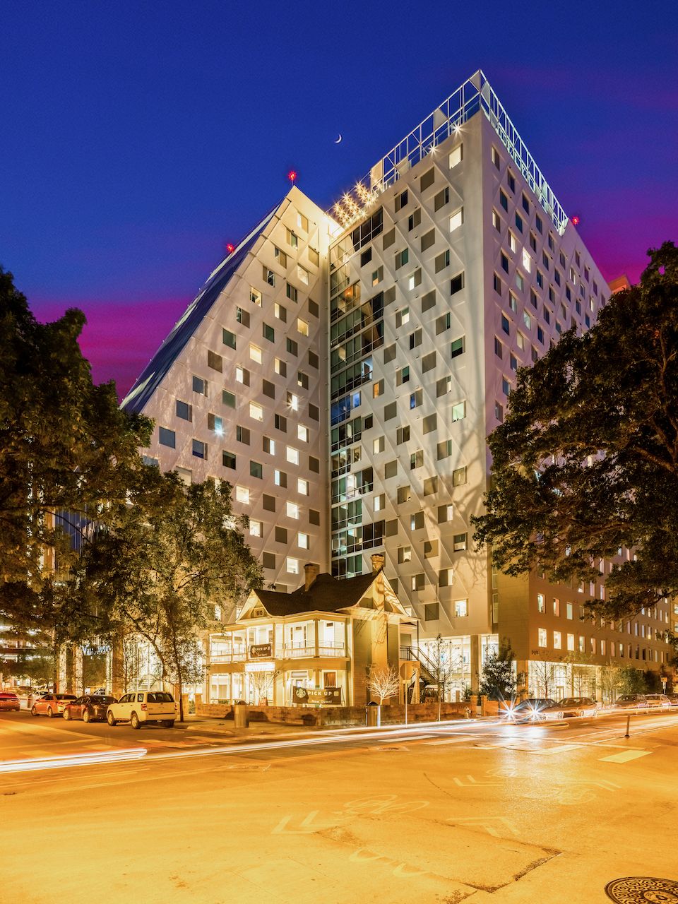 A modern high-rise building with a geometric facade, reminiscent of stylish UT apartments, towers over a small, older house at dusk. Bright streetlights and passing car light trails are visible in the foreground.